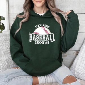 Könnte beinhalten: Ein dunkelgrüner Kapuzenpullover mit einem weißen Baseball-Motiv. Das Motiv enthält die Wörter "Team Name", "Baseball", "Sammy #9" und einen Baseball mit roten Nähten. Der Kapuzenpullover hat eine Vordertasche und eine Kapuze.