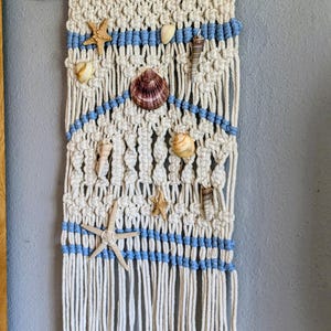 May include: Macrame wall hanging with a natural, off-white color scheme, accented with blue stripes and adorned with seashells and starfish. The wall decor is attached to a wooden dowel.
