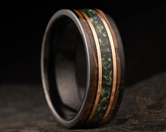May include: A close-up of a ring with a dark wood and metal band. The ring features a central inlay of green stone and is accented with thin gold-colored bands. The ring is set against a dark background.