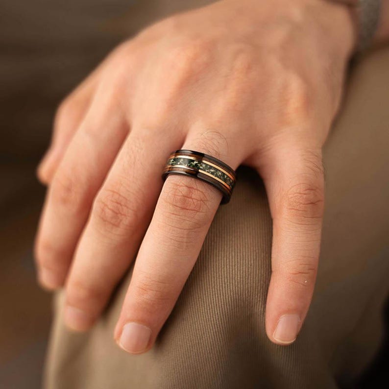 May include: A black ring featuring a green stone inlay and gold-coloured accents. The ring is displayed on a finger, highlighting its dark band and decorative details. The design includes a central green stone band and thin gold stripes.