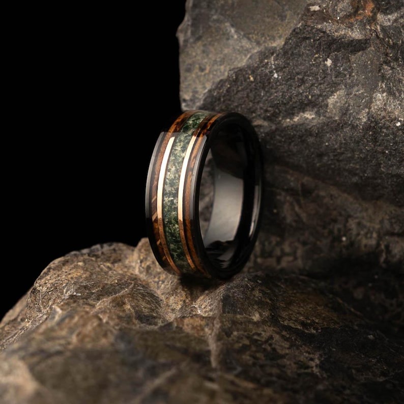 May include: A black ring featuring a central band of green stone, framed by bands of wood and gold-coloured metal. The ring is displayed against a dark backdrop and a textured rock, emphasising its design and materials.