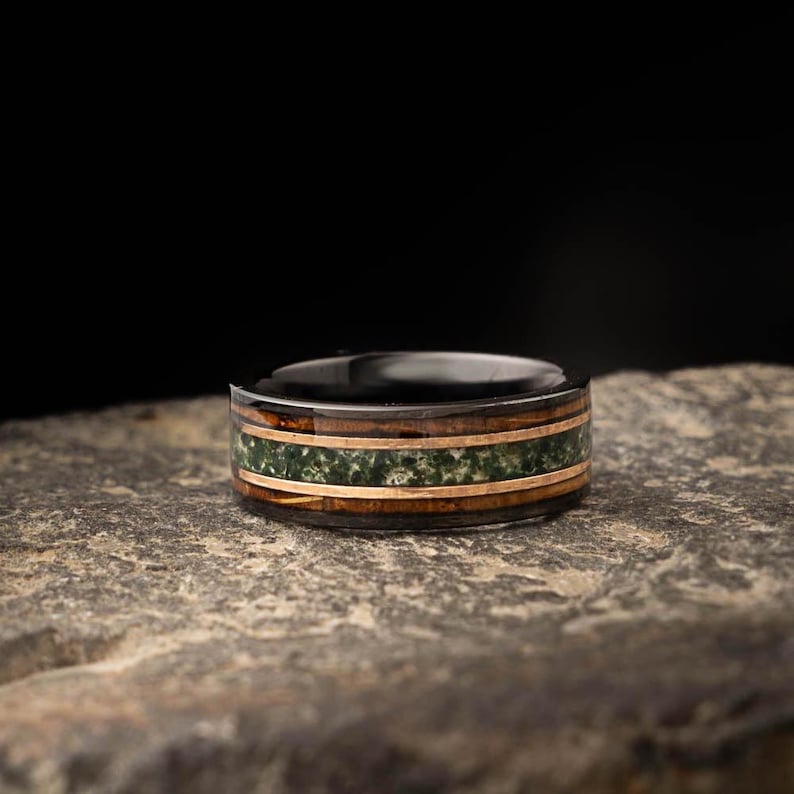 May include: A black ring featuring a wood grain band, a green stone inlay, and gold-coloured metal accents. The ring is displayed on a stone surface, highlighting its detailed design and craftsmanship.