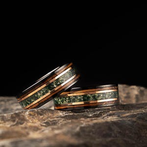 May include: Two wedding rings with a dark wood grain exterior, featuring a green stone inlay and thin copper bands. The rings are displayed on a textured, grey stone surface against a black background.