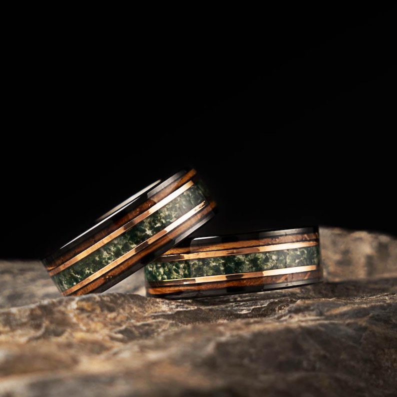 May include: Two wedding rings with a dark wood grain exterior, featuring a green stone inlay and thin copper bands. The rings are displayed on a textured, grey stone surface against a black background.