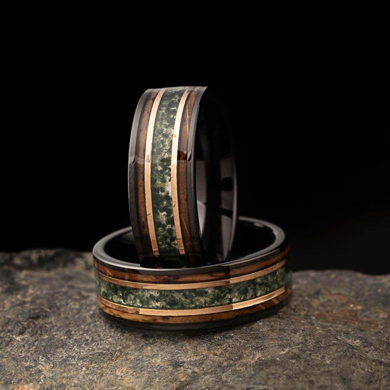 May include: Two rings with a black band, featuring a wood grain design with a green stone inlay and gold-coloured metal accents. The rings are stacked, highlighting the intricate details and craftsmanship.