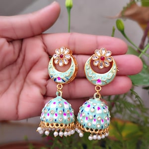 Puede incluir: Un par de pendientes dorados con un diseño floral. Los pendientes presentan una media luna de esmalte azul claro y una gota en forma de campana adornada con detalles blancos, rosas y azules. Pequeñas perlas blancas cuelgan de la parte inferior. Los pendientes están sostenidos por una mano.
