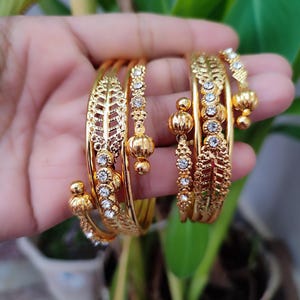 May include: Several gold bangles are held in a hand. The bangles have detailed leaf and floral patterns, with clear gemstones and hanging gold beads. The bangles are a bright, shiny gold colour.