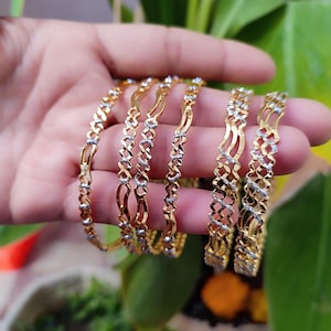 Könnte beinhalten: Sechs goldene Armreifen mit aufwendigen Designs und kleinen, funkelnden Akzenten. Die Armreifen werden in einer Hand gehalten, was ihre filigrane Handwerkskunst und ihr elegantes Aussehen hervorhebt. Der Schmuck ist vor einem verschwommenen Hintergrund aus Grün platziert.