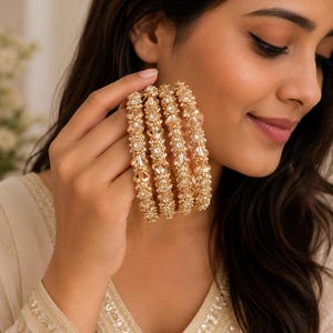 May include: A set of four gold-toned bangles featuring intricate metalwork and clear, faceted crystal beads arranged in a repeating pattern. The bangles have a metallic finish and a textured surface with small, sparkling stones embedded throughout.