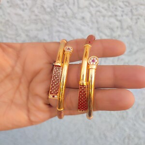 May include: Two gold bangles with red and clear detailing. Each bangle has a decorative red section with a textured pattern and a small, round, clear gem. The bangles are held in a hand.