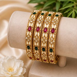 May include: A set of four gold-tone bangles featuring an off-white enamel inlay, accented with oval-shaped red and green stones, and small clear crystals arranged in floral patterns. The metal edges have a textured, rope-like design detail.