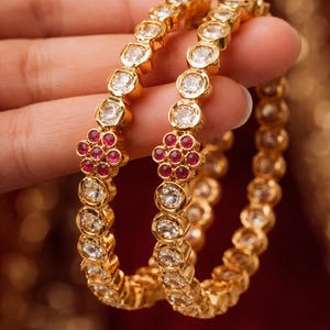 May include: Two gold bangles with clear and red gemstones. The bangles are made of gold and feature a repeating pattern of round, clear gemstones and clusters of red gemstones. The jewellery is displayed against a blurred red background.