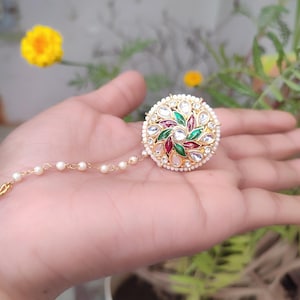 May include: A gold-coloured, round hair accessory with a floral design. The centre features red, green, and white gemstones. The accessory is surrounded by small white pearls and has a delicate chain of pearls.