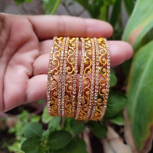 May include: Two gold bangles with detailed floral and scroll patterns. The bangles are embellished with small, clear, sparkling stones, giving them a luxurious and ornate look. The jewellery is shown against a background of green leaves.