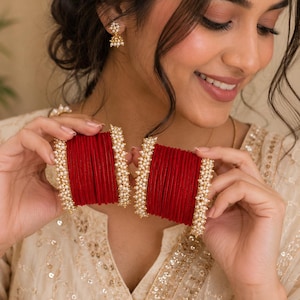 Peut inclure: Deux ensembles de bracelets joncs rouges empilés avec des bords en métal doré ornés de petites perles blanches. Les bracelets ont une texture mate et sont disposés en deux groupes distincts, chacun bordé de perles blanches délicates.
