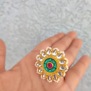 May include: A gold-coloured ring with a floral design. The ring features a central green circle with a red gem, surrounded by clear, petal-shaped stones and small white pearls. The ring is held in a hand.