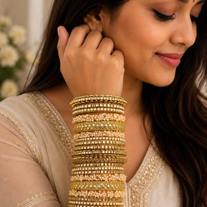 May include: A stack of gold-toned bangles featuring alternating patterns of small white beads and square-cut reflective mirror inlays. The set consists of multiple individual bracelets worn together to create a wide, textured, and metallic wrist ornament.
