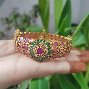 May include: A gold-coloured bangle bracelet set with oval-shaped ruby-coloured stones and smaller green stones. The bracelet has a central design with a larger ruby-coloured stone surrounded by green stones and clear accents. The jewellery is displayed against green foliage.