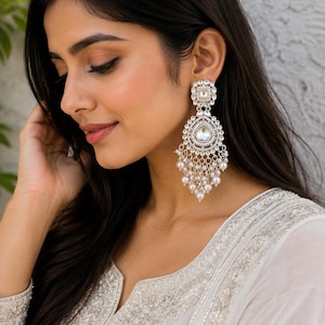 May include: A silver-toned chandelier earring featuring a square stud, a central teardrop-shaped clear stone, and a circular frame adorned with small white crystals and dangling white pearls arranged in a tiered, fringe-like pattern.