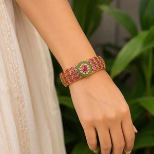 Könnte beinhalten: Ein goldfarbenes Armband mit drei Reihen ovaler rosa Steine. In der Mitte des Armbands befindet sich ein kreisförmiges Blumenmotiv mit einem zentralen rosa Stein, umgeben von kleinen grünen Steinen und winzigen, klaren weißen Akzenten.
