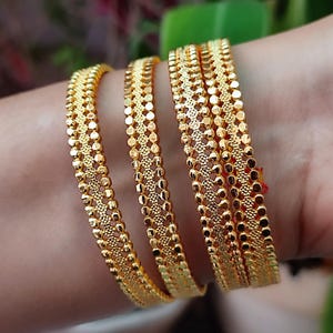 May include: Close-up of multiple gold bangles featuring detailed dot and floral patterns. The bangles are stacked on a wrist, displaying their intricate design and warm, golden hue. The background is blurred, focusing on the jewellery.