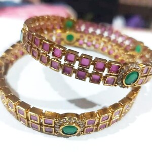 May include: Two gold bangles with pink and green gemstones. The bangles have a repeating pattern of square pink stones and are accented with larger green stones and small clear stones.