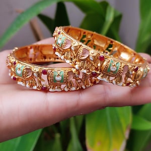 Könnte beinhalten: Zwei goldfarbene Armreifen mit aufwendigen Elefantenmotiven. Die Armreifen sind mit roten, weißen, rosa und grünen Emaille-Akzenten verziert. Die Elefantenfiguren sind detailliert, und die Armreifen haben eine strukturierte Oberfläche, die traditionelle Handwerkskunst zeigt.