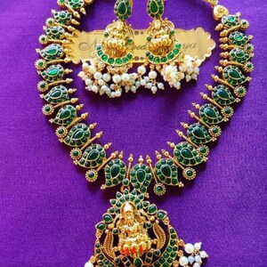 May include: A gold-toned necklace and earring set with green gemstone accents and white pearl clusters. The necklace features a pendant with a central figure. The earrings have a similar design. The jewelry is displayed on a purple surface.
