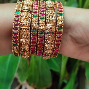 Puede incluir: Un conjunto de brazaletes dorados con diseños intrincados y piedras preciosas rojas y verdes incrustadas. Los brazaletes presentan figuras y patrones detallados, creando un aspecto tradicional y ornamentado. Las joyas se exhiben sobre un fondo verde borroso.