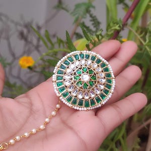 May include: A gold-coloured, circular headpiece with a central floral design. It features emerald green and clear gemstones, surrounded by small white pearls. A delicate chain of pearls extends from the headpiece.