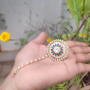 May include: A gold-coloured headpiece featuring a central design of red, green, and clear gemstones, encircled by small white pearls. A delicate strand of pearls extends from the headpiece, ending in a small loop. The headpiece is held in a hand.