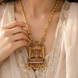Op de afbeelding: Een goudkleurige ketting met een lange schakelketting met bolvormige kralen en een grote, rechthoekige hanger. De hanger toont een centrale godheid zittend op een lotus, geflankeerd door twee staande figuren, met kleine parels aan de onderkant.