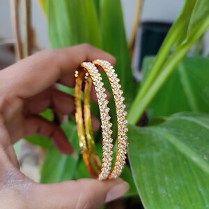 Può includere: Due braccialetti d'oro con un motivo scintillante a forma di diamante. I braccialetti sono tenuti davanti a uno sfondo di foglie verdi. I gioielli sono di un colore oro brillante e le pietre sono chiare.