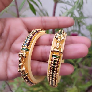 Op de afbeelding: Twee goudkleurige armbanden met ingewikkelde ontwerpen. De armbanden hebben een crèmekleurige basis met gouden accenten en zijn versierd met rode, groene en gouden details. Het ontwerp omvat een gedetailleerde olifantenkop en kleine, ronde versieringen.