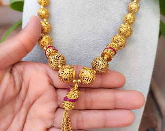 May include: A gold-colored necklace featuring a series of intricately designed, spherical beads. The necklace includes a tassel pendant with a cluster of small, dangling chains. The design incorporates small, red accents, adding a touch of color to the overall piece.