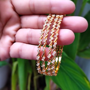 May include: Gold bangles with a zigzag pattern, embellished with small, sparkling gemstones. The bangles are held against a backdrop of green foliage, highlighting their intricate details and radiant shine. These elegant bangles are a beautiful accessory.