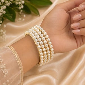 May include: A set of four stacked bangles featuring rows of small, round, cream-coloured pearls set against a gold-toned metal base. The bracelets are worn together on the wrist, creating a uniform, layered appearance with a smooth, polished finish.