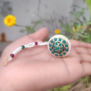 May include: A gold-coloured, round hair accessory featuring emerald-green square stones at its centre. It is encircled by small white beads and has a white string with red and green beads, and a gold hook.