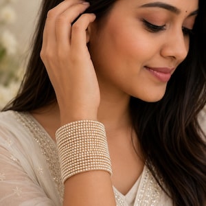 May include: A wide bangle bracelet composed of multiple thin, stacked bands. Each individual band features a continuous row of small, round, cream-coloured faux pearls set against a metallic gold-toned base, creating a textured, uniform appearance.