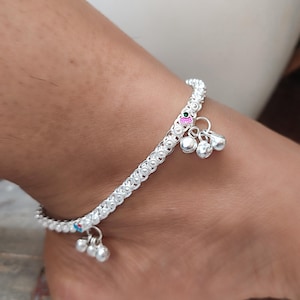 Puede incluir: Una tobillera plateada con un diseño floral y pequeñas campanas colgantes. La tobillera se lleva en el tobillo. Las campanas tienen una superficie brillante y reflectante. La tobillera tiene pequeños acentos de color.