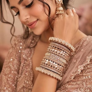 May include: A stack of gold-toned bangles featuring intricate metalwork, clear crystals, and small white pearls. The set includes various textures, including beaded edges and geometric crystal patterns, worn on the wrist alongside a matching jhumka earring.