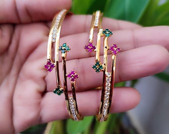 May include: Gold bangles with embedded clear and colourful gemstones. The bangles feature a row of clear stones and are accented with clusters of green and pink gemstones. The jewellery is held against a backdrop of green foliage.