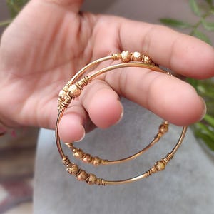 Könnte beinhalten: Zwei goldfarbene Armreifen mit dekorativen Perlenakzenten. Die Armreifen werden in einer Hand gehalten, wodurch ihr filigranes Design zur Geltung kommt. Die Perlen sind in Gruppen entlang der Bänder angeordnet und verleihen dem Schmuck Textur und visuelles Interesse.