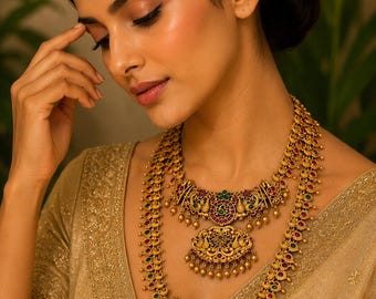 May include: A multi-layered gold-toned necklace featuring intricate metalwork, peacock motifs, and embedded red and green stones. The design includes two distinct pendants with dangling gold beads, suspended from chains adorned with repeating floral patterns.