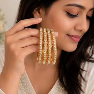 May include: A set of four gold-toned bangles featuring a continuous row of clear, round-cut stones set along the outer surface. Each bangle has a polished metallic finish and a uniform circular shape, designed to be worn as a stackable jewellery set.