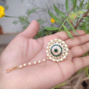 May include: A gold-coloured headpiece with a central green gem surrounded by white pearls. The circular design features a floral pattern. A delicate chain of small pearls extends to a hook. The headpiece is held in a hand against a blurred green background.