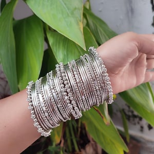 Op de afbeelding: Een pols versierd met een verzameling zilveren armbanden. De armbanden tonen een reeks ontwerpen, sommige met een gladde, gepolijste afwerking en andere versierd met kleine, kralen details. De armbanden zijn gestapeld, waardoor een gelaagd effect ontstaat.