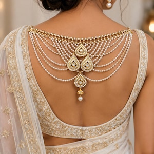 May include: A gold-toned back necklace featuring multiple draped strands of small white pearls and four teardrop-shaped pendants encrusted with clear crystals. The jewellery piece is worn over a white sleeveless garment with gold embroidery.