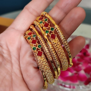 Könnte beinhalten: Ein Satz goldener Armreifen mit aufwendigen Designs und roten und grünen Edelsteinakzenten. Die Armreifen werden in einer Hand gehalten, um ihre verzierten Details und ihr poliertes Finish zu präsentieren. Die Edelsteine sind quadratisch und rund.