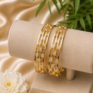May include: Two gold-toned bangles featuring a triple-band design connected by small vertical bars. Each band is set with multiple clear, round-cut stones that sparkle along the entire circumference of the circular jewellery pieces.